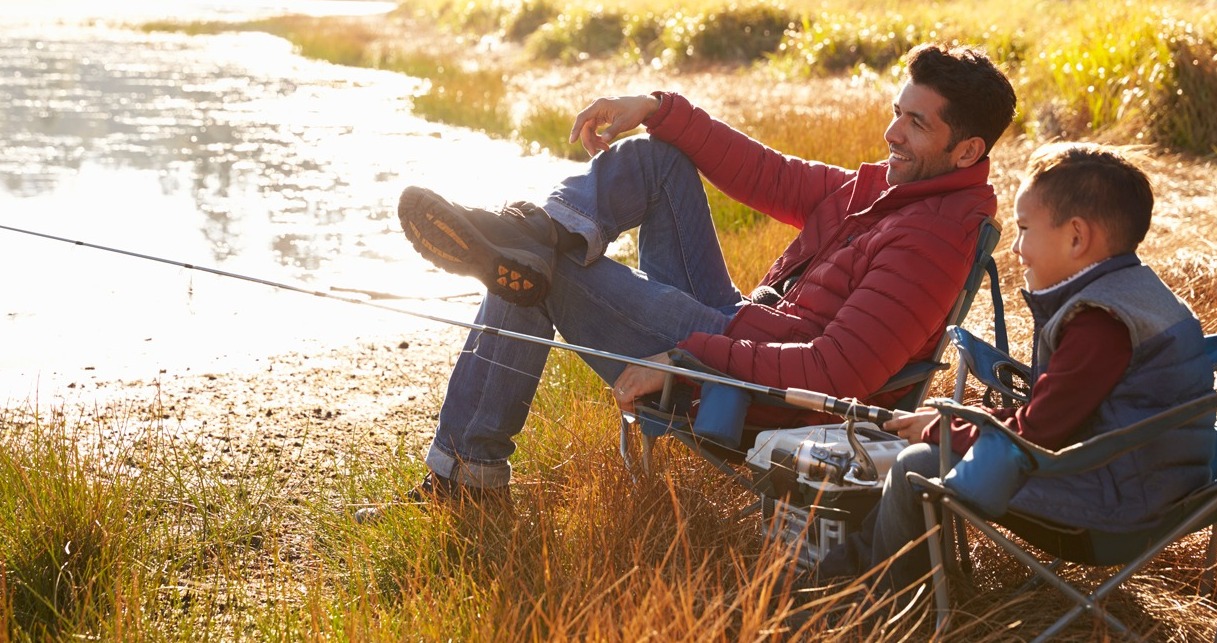 A father and his young son sit in folding chairs along the grassy bank of a river while the son casts out his fishing rod.